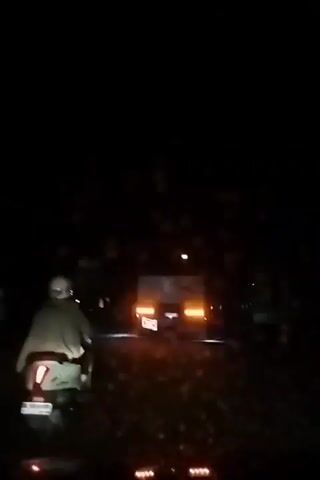 A nighttime video shows a person on a scooter in the foreground, moving on a road. Behind the scooter, a truck with its brake lights illuminated is visible. The truck appears to be moving away from the camera. The road is wet, suggesting it might be raining, as indicated by small circles of light scattered across the screen, resembling raindrops on a lens or windshield. The environment is dark, with only the lights from the vehicles and a distant street lamp providing illumination. The video is shaky, possibly due to the movement of the scooter.
