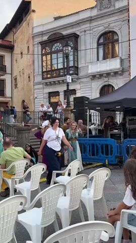 A group of people, some in historical costumes, are gathered in an outdoor square. White plastic chairs are arranged in rows, facing a stage area where musical equipment and a tent are set up. People are milling about, some standing and talking, others seated. One woman in a blue and green dress is walking towards the stage. Another woman in a purple dress and head covering is talking to a person in a black habit. A man in a green shirt and yellow pants is seated in one of the chairs. Buildings with balconies and windows surround the square. The overall impression is of a community event or festival.