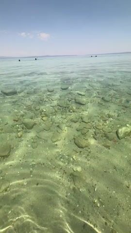 This is a video of a clear, shallow body of water, likely the sea given the location context. The seabed is visible and consists of sand and numerous smooth, rounded stones of varying sizes. Sunlight filters through the water, creating rippling patterns on the sandy bottom. In the distance, the surface of the water extends to a hazy horizon, with faint outlines of land visible. The sky is a clear, bright blue with a few wispy clouds. There are several small, dark shapes on the surface of the water in the distance, which appear to be people in small boats or on watercraft, or possibly swimmers. The overall impression is one of a calm, sunny day at a beach or shallow coastal area. The lighting suggests it is daytime, likely mid-morning or afternoon. There is no visible text in the video.