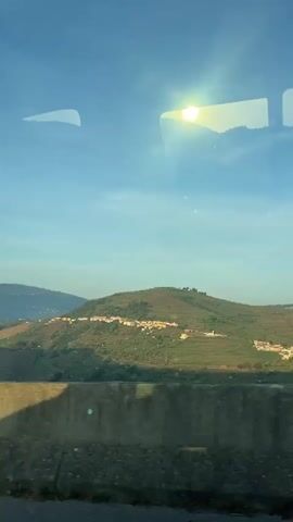 The video shows a moving landscape of rolling hills under a clear blue sky, viewed from inside a vehicle. The bright sun is visible, shining through the window. In the distance, the hills are covered with green terraced fields and small villages. A concrete barrier is seen in the foreground, partially obstructing the view. The shadow of an object, possibly from the vehicle, is cast on the barrier.