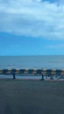 The video shows a view from a moving vehicle. The foreground is a dark, textured surface, likely asphalt. Behind this is a strip of ground with some dry grass and small stones. A metal railing with yellow and black stripes runs horizontally across the middle of the frame. Beyond the railing is the ocean, with gentle waves rolling towards a sandy or rocky shore. The water is a muted blue-gray. The sky above is a bright blue with scattered white clouds. The camera perspective suggests the vehicle is traveling parallel to the coastline. There are no people or animals visible. The overall impression is of a coastal drive on a slightly overcast day.
