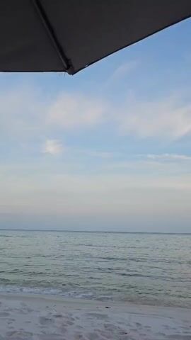 The video shows a close-up of a person's bare feet crossed at the ankles, resting on a black surface, presumably a lounge chair. The feet are positioned in the foreground. Beyond the feet, a white sandy beach stretches out. Gentle waves lap at the shore of a calm, blue ocean. In the distance, a faint horizon line separates the sea from a pale blue sky dotted with wispy white clouds. The top left corner of the frame shows a portion of a dark gray umbrella, providing shade over the scene. The overall impression is one of relaxation on a beach.