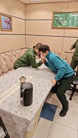 Three men are seated around a table engaging in arm wrestling, while a fourth man stands nearby observing and using a mobile phone. The individuals around the table appear to be of Asian descent. The two arm wrestlers, one in a green military-style uniform and the other in a blue shirt, are focused intensely on the competition. One man is positioned along the sofa, appearing to use it as leverage. The fourth man, also in a green military-style uniform, stands to the side, looking at his phone. The scene takes place in an enclosed room decorated with framed artwork on the walls, featuring natural and wildlife scenes. The table is covered with a patterned tablecloth, and an electric kettle sits prominently on the tabletop.