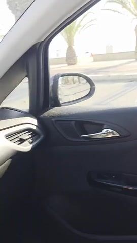 The video is captured from inside a vehicle, looking out through the driver's side window and then the windshield. The scene is a sunny day with a clear blue sky. Palm trees line a street or promenade, indicating a warm climate, possibly coastal. In the foreground, the interior of the car is visible, including the door panel, handle, and a portion of the dashboard. Outside, there is a paved area that appears to be a parking lot or sidewalk, with a few parked cars visible further down the road. A sign with a blue 'P' indicating parking is also present. The sea is visible in the distance beyond the palm trees. The car appears to be moving slowly or is stationary.