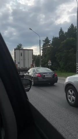 The video shows a car in traffic on a highway. The camera is positioned inside a car, looking out the driver's side window. Initially, a dark-colored Nissan Teana is visible in the adjacent lane, slightly ahead of the camera car. A large white truck with a Schmitz Cargobull logo is directly in front of both vehicles. The truck has "R" marked on its back. As the video progresses, the camera car advances, and the Nissan Teana moves out of view. The focus shifts to the white truck and the traffic ahead, including a blue car visible in the distance. The sky is cloudy. The interior of the camera car is partially visible, including the rearview mirror and the car door frame.