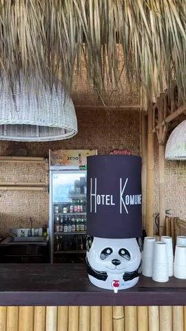 The video shows a close-up view of a water dispenser designed like a panda, situated on a wooden bar. The dispenser has a black cylinder on top with "HOTEL KOMUNE" written in white lettering. To the left of the dispenser, there's a refrigerator filled with bottles of beverages, and behind it, a counter with some cups. To the right of the dispenser, a stack of white paper cups is visible. The entire scene is under a thatched roof, suggesting an outdoor or tropical setting. The camera remains stationary throughout the short clip.
