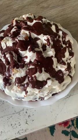 A cake sits on a white plate on a table. The cake is covered in white cream and drizzled with chocolate sauce. The cake appears to be made of cake pieces mixed with cream. The camera slowly zooms out and pans around the cake. The cake is on a wooden table. A colorful rug is visible in the lower part of the frame.