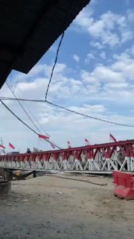 The video shows a low angle shot of a red and white bridge, likely in Indonesia judging by the Indonesian flags lining the bridge, under a partially cloudy blue sky. The camera seems to be located under a partially visible metal roof or awning, and there are power lines visible in the foreground. The area around the bridge appears to be unpaved with visible soil and some construction barriers. People are walking and cycling on the bridge.
