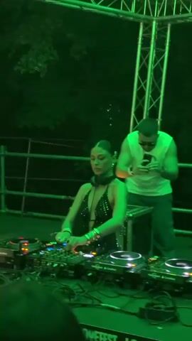 A woman wearing a sparkly black halter-neck top and headphones is seated at a DJ booth, her hands on the equipment. Behind her, a man in a white sleeveless shirt and sunglasses stands, looking at his phone. The DJ booth has multiple pieces of electronic equipment, including turntables and mixers, with wires visible. The scene is illuminated with green lighting, creating a stage-like atmosphere. Metal scaffolding and trees are visible in the background. The word "LOVEFEST" is visible on the front of the DJ booth.