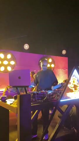 A man wearing headphones and a dark shirt with green accents is standing behind a DJ setup, which includes a laptop and various electronic equipment. He is illuminated by bright yellow and pink stage lights, and there is some smoke or haze in the air. To the left of the DJ, a woman in a silver, flowing garment is dancing energetically, with her arms raised. In the background, there is a wall with circular light fixtures and a couch on the far left. The scene appears to be a live performance or event in a dimly lit venue.