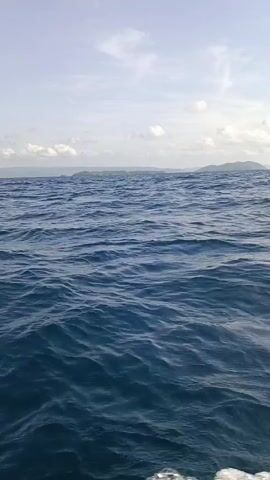 The video shows a wide shot of the ocean with choppy, dark blue waves. The horizon is visible with a faint line of islands in the distance. The sky above is a pale blue with scattered white clouds. The camera appears to be positioned on a moving boat, as evidenced by the foamy white wake at the bottom of the frame. There are no people or other objects visible in the scene.