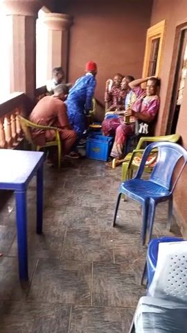 The video shows several African men sitting on a tiled porch area. Some are seated on plastic chairs, while others are sitting against the wall. One man is standing and has a scarf draped over his arm. He is wearing a blue tunic and a red hat. There is a blue plastic crate in the middle of the group, and some beer bottles and food containers are visible. One of the men is laughing, while another has a beer bottle in his hand. A small table and a door are also visible in the scene.