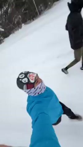 The video shows two people in a snowy environment. One person, wearing a bright blue jacket and a patterned hat and scarf, appears to be filming while sliding or being pulled on the snow. This person is close to the camera, giving a first-person perspective. The second person, dressed in a black jacket and olive pants, is running alongside or slightly ahead of the first person on the snow-covered ground. The background shows a snowy landscape with trees lining the perimeter. Later, the video shows a snowy path with tracks and a wooden building in the background. The focus remains on the snowy terrain and surrounding environment.