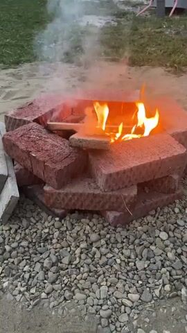 The media shows a close-up view of a DIY fire pit constructed from red bricks. The fire pit is hexagonal in shape and sits on a bed of gravel. Inside the fire pit, a small fire is burning, with visible flames and smoke rising. Several pieces of wood are burning, and one piece appears to be a plank of wood. The fire pit is surrounded by gravel and sand. In the background, there is green grass, and hints of structures and possibly a playground can be seen. The overall setting appears to be outdoors, likely a backyard or a recreational area. The time of day is unclear, but the lighting suggests it might be dusk or late afternoon. There is no visible text in the media file. The scene evokes a sense of relaxation and outdoor leisure, perhaps a small gathering or a quiet moment by the fire.