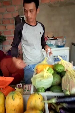 A man in a grey long-sleeved shirt and blue jeans stands behind a market stall, arranging produce. In front of him are baskets filled with papayas, avocados, squash, chayote, and eggplants. To his right, a basket of green chili peppers is visible. A woman in a red headscarf and shirt is partially visible behind the man, near a scale. In the background, shelves are stocked with more vegetables, and a white refrigerator or cooler is present. The stall appears to be indoors, with a brick wall as the backdrop. The man is holding a plastic bag and interacting with the produce.