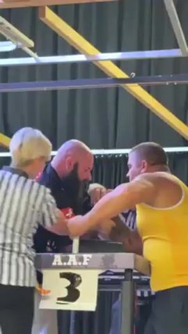 Two men are arm wrestling. A woman in a referee uniform observes them. The man on the left has a long beard and a black shirt. The man on the right has on a yellow tank top. They are competing at table number 3. The walls are dark, but light is coming from above.