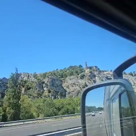 The video begins with a view of a road and a hill with a building on top. The camera appears to be inside a vehicle, possibly a truck, as a side mirror is visible on the right side of the frame. The sky is clear and blue. As the video progresses, the vehicle moves, and the view of the hill and the building becomes clearer. The building on the hill appears to be a church or a similar structure with a tall spire. The hill is covered with trees and rocks. In the side mirror, other vehicles can be seen on the road, indicating that this is likely a highway or a busy road. The overall scene suggests a journey through a scenic area, possibly in the countryside or a mountainous region.