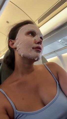 A woman is on an airplane wearing a white facial mask. She wears a blue tank top. A child's face appears in the corner of the frame. An airplane speaker message is audible.