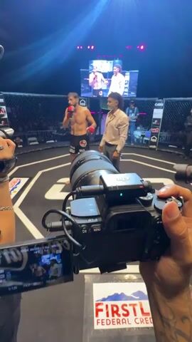 A fighter stands in a martial arts octagon, facing a man in a light-colored shirt. A camera is pointed at the fighter from the foreground. Another camera operator is visible in the background, holding a camera. A large screen displays images of people. The octagon is lit with spotlights. A banner for "First Federal Credit Union" is visible on the floor.
