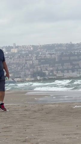 The video depicts an overcast day at the beach with a cityscape in the background. An older man wearing a dark polo shirt, gray shorts, a white cap, and sunglasses walks towards the camera, holding a water bottle. The beach is sandy with visible debris and waves crashing on the shore. After the man passes, the camera focuses on the beach and city view, revealing other individuals walking along the shoreline. The city in the background is dense with buildings on a hillside, obscured by the cloudy weather.