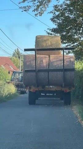 A tractor with a front loader is partially visible on the left side of a paved road. The tractor is positioned near a ditch and some vegetation. The main focus of the video is a large trailer being pulled by an unseen vehicle, likely another tractor or truck, which is moving away from the camera. The trailer is loaded with numerous large, round hay bales. The hay bales are stacked two high and appear to be secured to the trailer with metal bars. The trailer has visible tires and a license plate. The scene takes place on a sunny day with a clear blue sky. Trees and houses are visible in the background, suggesting a rural or semi-rural setting. Electrical wires are strung overhead. The camera appears to be positioned inside a vehicle, looking forward through a windshield.