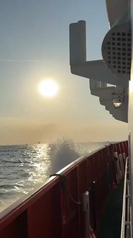 A boat is moving through the water at sunset. The sun is low in the sky, casting a warm glow over the ocean. The boat is leaving a trail of spray in its wake. The water is choppy, and waves are crashing against the side of the boat. The sky is clear, with a few wispy clouds. The scene is peaceful and serene.