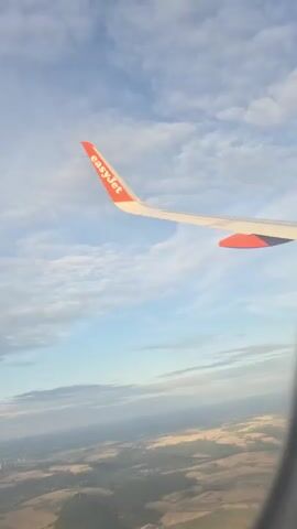 A view from an airplane window shows the wing of an easyJet airplane extending into a blue sky with scattered clouds. Below the wing, a landscape of rolling hills and fields is visible, with a small town or village nestled in the lower left portion of the frame. The sunlight suggests it is either morning or evening. The video is short, with no discernible action or interaction beyond the passive observation from the aircraft.