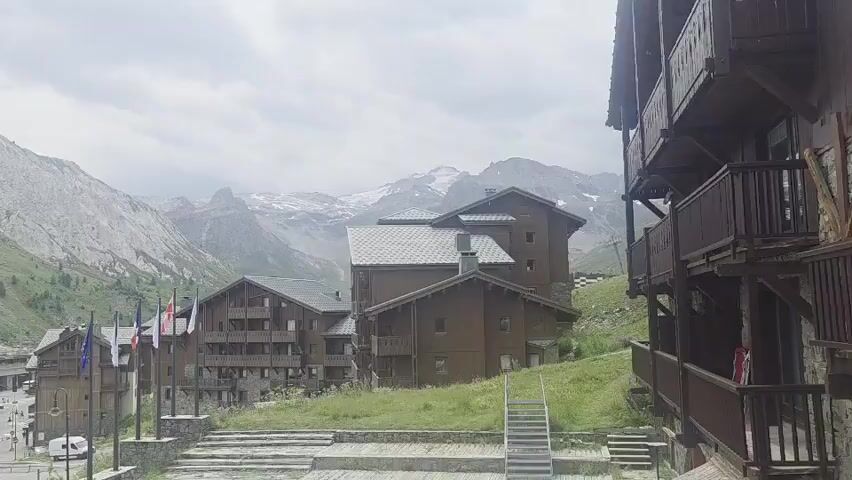 The video shows a wide shot of a mountain village on an overcast day. In the foreground, there is a wooden building with balconies. To the left, there are several flags on poles, including the French flag. Behind the flags is a street with some parked cars and a white van. Further into the background, there are more buildings, mostly made of wood, nestled into the side of a green, mountainous landscape. The mountains in the distance are tall and rugged, with some patches of snow visible on the highest peaks. The sky is cloudy and grey.
