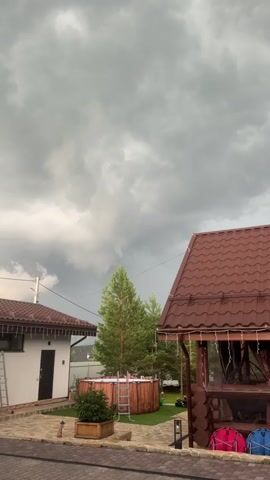 The video shows a backyard scene with a cloudy sky. On the left, there is a white building with a brown roof and a ladder leaning against it. In the center, there is a round wooden hot tub with a ladder. Behind the hot tub, there are pine trees. On the right, there is a brown wooden gazebo with a brown roof and string lights. Inside the gazebo, there are two colorful cushions, one pink and one blue. The ground is paved with bricks. The video appears to be taken from a slightly elevated position, looking down into the backyard. The clouds are dark and appear to be heavy, suggesting impending rain.