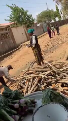 A man and a woman interact on a dirt road. The woman, wearing a green head covering and a patterned skirt, is handing something to the man, who is standing next to a motorcycle. A pile of firewood lies between them. In the foreground, a tray holds bundles of green onions. In the background, other women are walking down the road. Buildings and trees line the street, and the sky is clear.