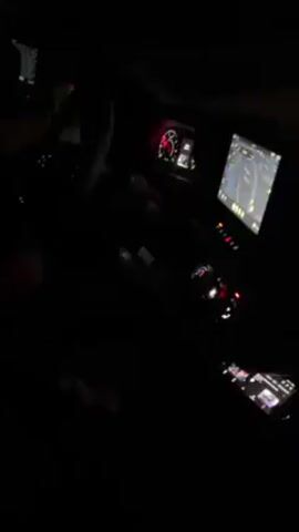 Here is a description of the video:

The video shows the interior of a vehicle at night. It is dark, but the dashboard lights are visible, illuminating the steering wheel, instrument panel, and a large touchscreen display showing a navigation map. A cell phone with its screen on is visible in the center console. The driver's hands can be seen on the steering wheel. The passenger seat is partially visible, appearing to be empty. The video seems to be taken while the vehicle is moving. There is audio of music playing and someone speaking in the background.