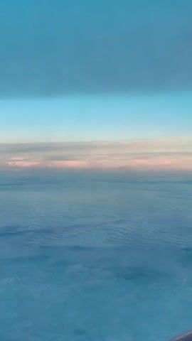 A video captured from an airplane window shows the wing of a SpiceJet aircraft against a backdrop of clouds and a twilight sky. The wing is positioned on the right side of the frame, angled upwards and towards the viewer. The sky transitions from a deep blue at the top to lighter shades of blue and pink near the horizon. Below the wing, a vast expanse of white clouds stretches out, appearing soft and textured. The lighting suggests either dawn or dusk. No people are visible in the shot. The overall scene evokes a sense of flight and the serene beauty of the upper atmosphere.
