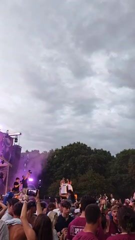 A vertical, low-angle shot captures an outdoor music festival under a cloudy sky. The stage on the left is adorned with purple lights and large speakers. Two performers are visible on the stage, one holding a microphone and the other gesturing. Smoke drifts from the stage area. In the center of the frame, two individuals stand on the shoulders of others in the crowd, raising their arms. A dense crowd of spectators fills the foreground, many holding up phones to record the performance. The overall atmosphere appears energetic and celebratory.