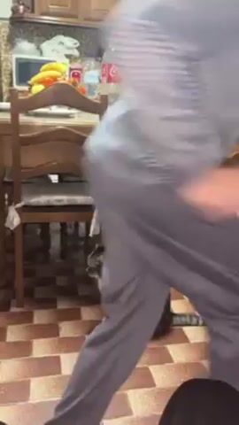 The video shows a kitchen scene with a young boy dancing near a tabby cat. The boy, wearing a blue patterned long-sleeved shirt and gray pants, enters the frame and dances with a playful expression. The cat sits on the checkered tile floor near a wooden table and chairs. The kitchen has wooden cabinets, a countertop with various items including fruit and a Coca-Cola bottle, and an oven.