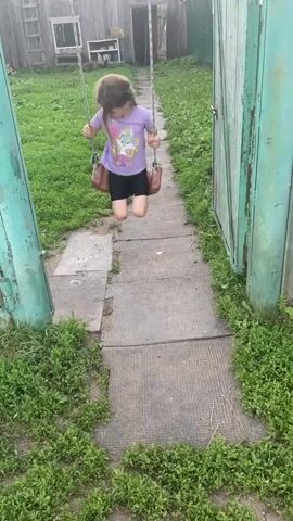 A young girl with light brown hair is swinging on a swing set in an outdoor yard. She is wearing a purple t-shirt with a graphic of a rabbit and the words "what's up!" and "RISING STAR" printed on it. She is also wearing black shorts, white socks, and black crocs. The swing set is made of metal and rope, with a brown plastic seat. The yard is covered in green grass and has a concrete path leading away from the swing set. In the background, there is a wooden fence, a shed, and a ladder leaning against a building. The lighting suggests it is daytime, possibly overcast given the diffused light. The girl appears to be enjoying herself, with a slight smile on her face. The overall scene suggests a casual, rural setting.