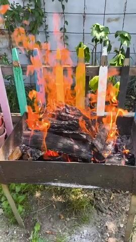 A close-up shot focuses on a metal brazier filled with burning logs. The fire is bright orange and yellow, with some smoke rising. Behind the brazier, a wooden fence is visible, decorated with colorful, oversized pencil-shaped posts. These posts are painted in various bright colors, including green, blue, yellow, and white, with some showing black tips resembling pencil points. The background consists of green foliage and a light-colored wall or fence, suggesting an outdoor setting. The overall impression is a rustic and somewhat whimsical scene.