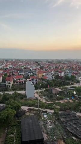 This is a drone video of a rural village in Vietnam during sunset. The camera pans slowly across the landscape, revealing a densely packed village with red-tiled roofs and a few taller buildings in the distance. The sky is a soft gradient of orange and pink, indicating the sun is setting. The foreground shows agricultural fields with black netting covering some of the crops. The overall atmosphere is peaceful and serene.