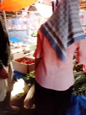 The video shows a market scene. In the foreground, a person wearing a pink and black polka dot shirt and a headscarf is partially visible. In the middle ground, a person wearing a dark jacket and light-colored pants is standing next to a wooden crate filled with red tomatoes. Behind them, a stall is laden with various items, including what appear to be toys and clothing accessories. Another person in a yellow shirt is visible further back, near a blue tarp. The market appears to be outdoors, with an orange tarp overhead providing shade. The lighting is dim, suggesting it might be evening or an overcast day. The overall impression is a busy marketplace.