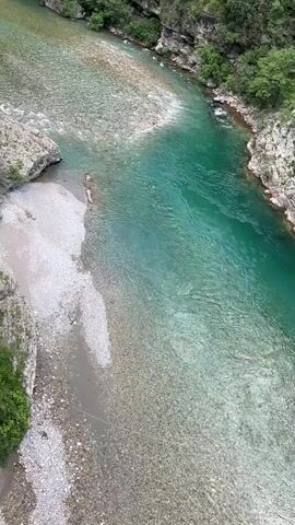 The camera pans up from an overhead view of a turquoise river to the top of a lush mountain. The river has stony banks and large boulders alongside green trees and bushes. The mountain has a rocky surface with some green trees and bushes. The sky above the mountain is partly cloudy and blue.