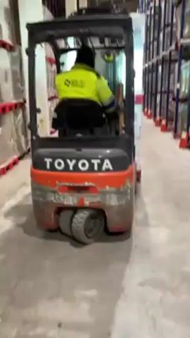 The video shows a forklift being driven in a warehouse. The forklift is a Toyota, orange and grey. The operator is wearing a bright yellow vest with the word "SBLOG" on the back and a hat or hood. The forklift is carrying a pallet stacked with boxes shrink-wrapped in plastic. The warehouse is tall with rows of shelves stocked with goods.