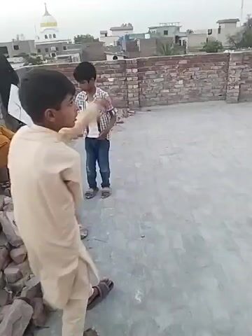 The video shows two children on a rooftop. One child, wearing a light-colored outfit, is facing away from the camera, gesturing with his arm. The other child stands in the background, wearing a plaid shirt and jeans. The camera then pans up to show a kite flying in the sky against a gray, overcast sky. The rooftops of nearby buildings are visible in the background. The rooftop the children are standing on has a low brick wall. Some loose bricks are piled on one side.