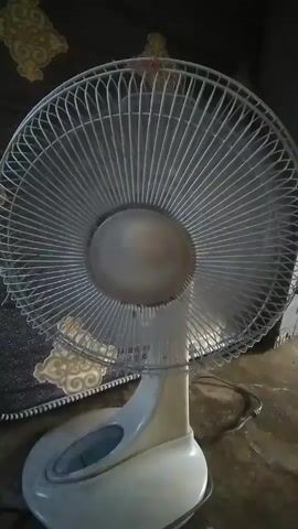 The video shows a close-up view of a white oscillating fan with a metal grill. The fan is turned on and the blades are spinning, creating a visible blur. The fan is positioned on a surface that appears to be a floor, with a patterned rug or fabric in the background. The lighting is dim.