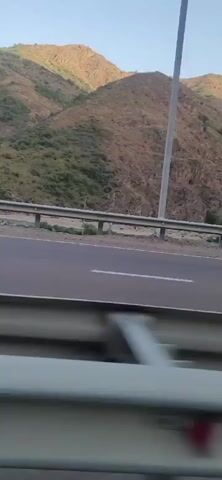 The video shows a view from a moving vehicle on a highway that runs alongside a steep, mountainous terrain. The mountains are covered in sparse vegetation, with areas of rocky, exposed slopes. The sky is clear and blue, indicating daylight. A metal guardrail separates the highway from the mountain. The road has white lane markings. The camera is positioned slightly downwards, capturing the road and the guardrail in the foreground and the mountains in the background. The footage appears to be taken during the daytime.