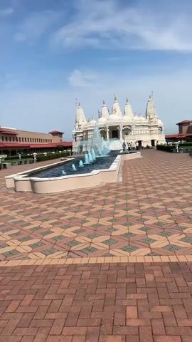 A wide-angle shot captures the exterior of a magnificent white Hindu temple with multiple spires. In the foreground, a decorative fountain sprays water, with some jets illuminated in a bright blue hue. The fountain is set in a dark-colored pool, bordered by a light-colored rim. The ground leading up to the temple and surrounding the fountain is paved with a patterned arrangement of reddish-brown and tan bricks.

To the left of the temple, a long, multi-story building with a terracotta-colored roof is visible. The sky above is a clear blue with scattered white clouds, suggesting a pleasant day. A few people are walking around the temple grounds, including near the main entrance. The overall scene conveys a sense of grandeur and tranquility, characteristic of a religious and cultural site. The architecture of the temple is intricate, with detailed carvings and ornamentation adorning its facade. The time of day appears to be daytime, given the bright sunlight and clear sky. The location cues point to Bartlett, United States of America, implying this is a significant religious structure within that region.