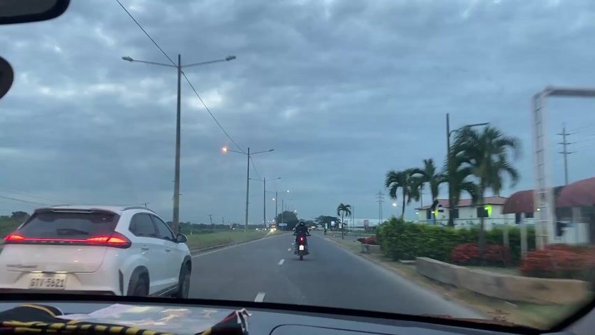 The view from inside a car driving on a two-lane road. A motorcycle is ahead, traveling in the same direction. A white SUV is briefly visible to the left. Streetlights illuminate the road under a cloudy sky. Palm trees and a building are visible on the right side of the road. A large truck appears on the left side of the road, moving in the opposite direction. Power lines and poles stretch across the sky. A bus stop and a billboard are visible further down the road on the right.