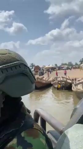 The video shows a group of soldiers riding in a wooden boat on a river. The soldiers are wearing helmets and camouflage uniforms. In the background, there is a riverbank with people and boats. The sky is blue with white clouds. One soldier is standing on the boat, looking out at the water. The boat is moving towards the riverbank.