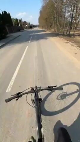 The video shows a first-person perspective of someone riding a bicycle on a paved road. The road has a white line dividing the lanes, and there are trees on one side and houses on the other. The sky is blue, and the sun is shining, casting a shadow of the bicycle and the rider's leg on the road.  A person is seen walking in the distance ahead.