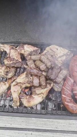 The video shows a close-up, overhead view of a grill filled with various meats cooking over hot coals. The meats include several pieces of chicken, appearing to be legs and thighs, some browning and slightly charred. Interspersed with the chicken are rows of sausages, some appearing to be skinless, elongated meatballs or mince, and also some larger, reddish-brown sausages which are likely a different type of sausage. Smoke is rising from the grill, indicating that the food is actively cooking. The grill itself is made of metal bars and sits on a wooden surface. The background is dark and out of focus.