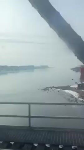 The video shows a view from a moving vehicle, likely a train, crossing a bridge or viaduct. The exterior scene features a body of water with a coastline. The ground is covered in snow. In the distance, there's an industrial area with cranes and tall structures, possibly related to shipbuilding or offshore operations. The sky is overcast. The structural elements of the bridge or viaduct are visible in the foreground. No people are visible.