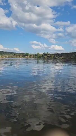 This is a vertical, first-person view video from a body of water, likely a river. The water is calm with ripples and reflections of the sky and clouds. In the distance, across the water, there is a sandy bank with sparse vegetation and some trees. On the bank, a group of people are visible, appearing to be sunbathing or socializing. The sky is blue with fluffy white clouds. The video appears to be taken on a sunny day.
